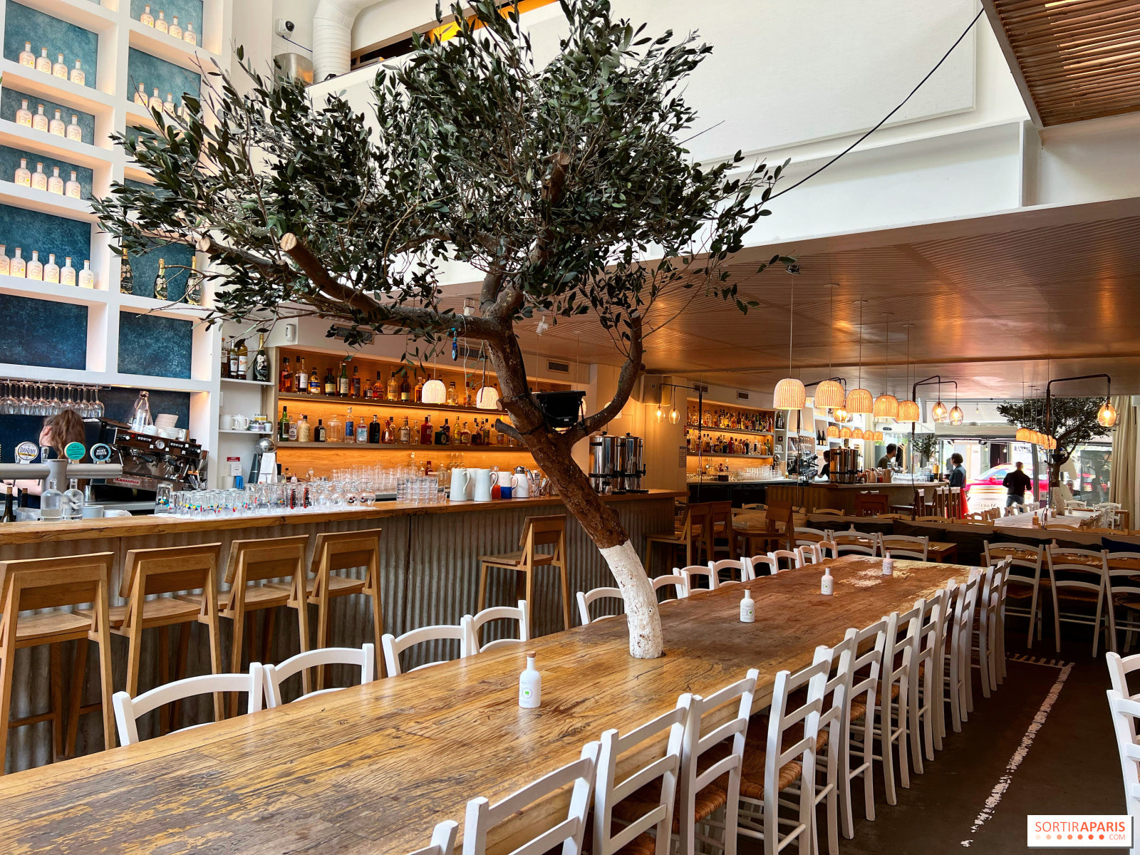 Intérieur du restaurant Yaya Secrétan à Paris, avec un grand bar en bois, une longue table conviviale entourée de chaises blanches et des luminaires modernes. Ambiance chaleureuse et méditerranéenne, lieu partenaire des soirées Crush Party pour des rencontres authentiques et festives.