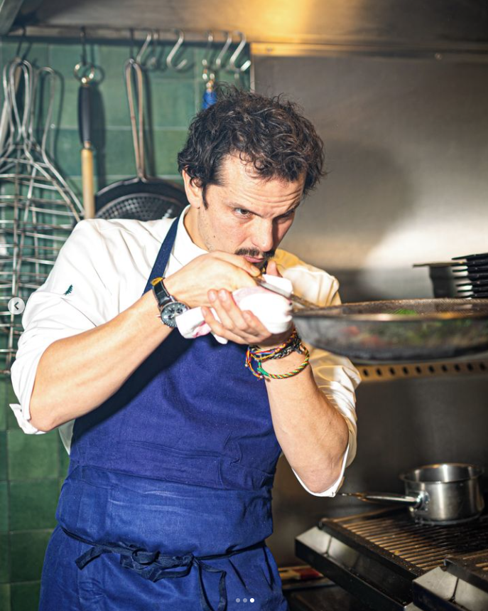 Chef cuisinier Juan Arbelaez
 concentré en train de préparer un plat dans une cuisine professionnelle, vêtu d'un tablier bleu. Ambiance chaleureuse et authentique, illustrant le savoir-faire culinaire des partenaires des soirées Crush Party, comme le restaurant Bazurto Paris.