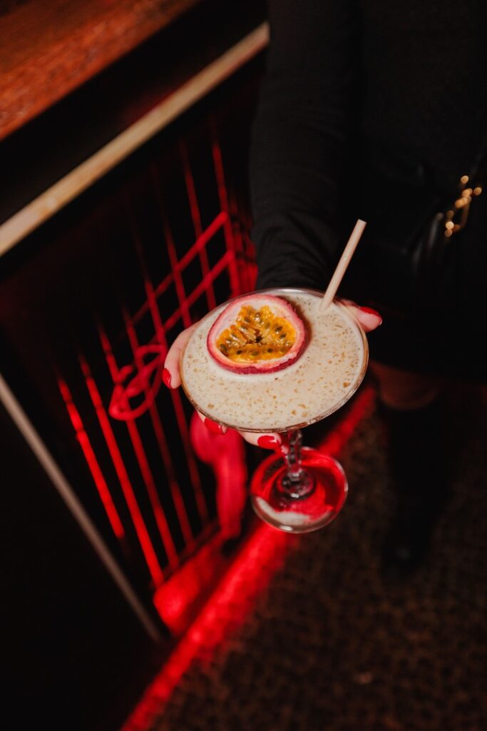 Photo d'un cocktail exotique garni d'un demi-fruit de la passion, tenu par une main élégante dans une ambiance tamisée. Capturée lors d'une soirée Crush Party, cette image met en avant le raffinement et la convivialité des événements parisiens dédiés aux célibataires. Un décor lumineux rouge et une présentation soignée ajoutent une touche festive et chic.