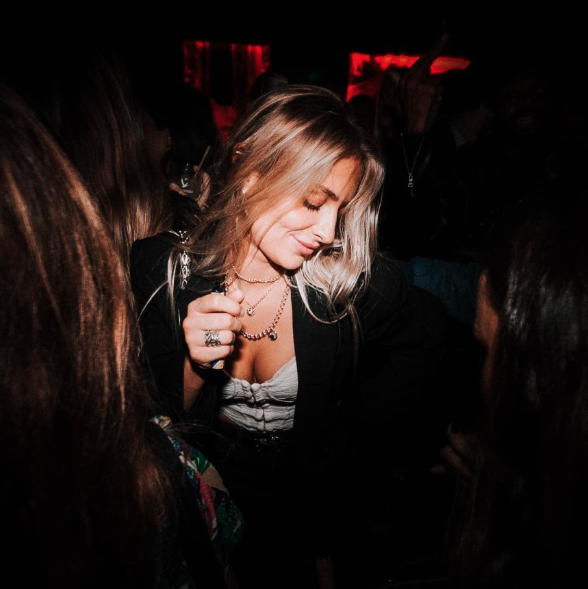 Photo d'une jeune femme blonde dansant avec élégance lors d'une soirée Crush Party. Habillée d'une tenue chic avec des accessoires raffinés, elle sourit subtilement dans une ambiance tamisée et festive. L'éclairage met en valeur l'énergie conviviale et authentique de l'événement, dédié aux célibataires à Paris.