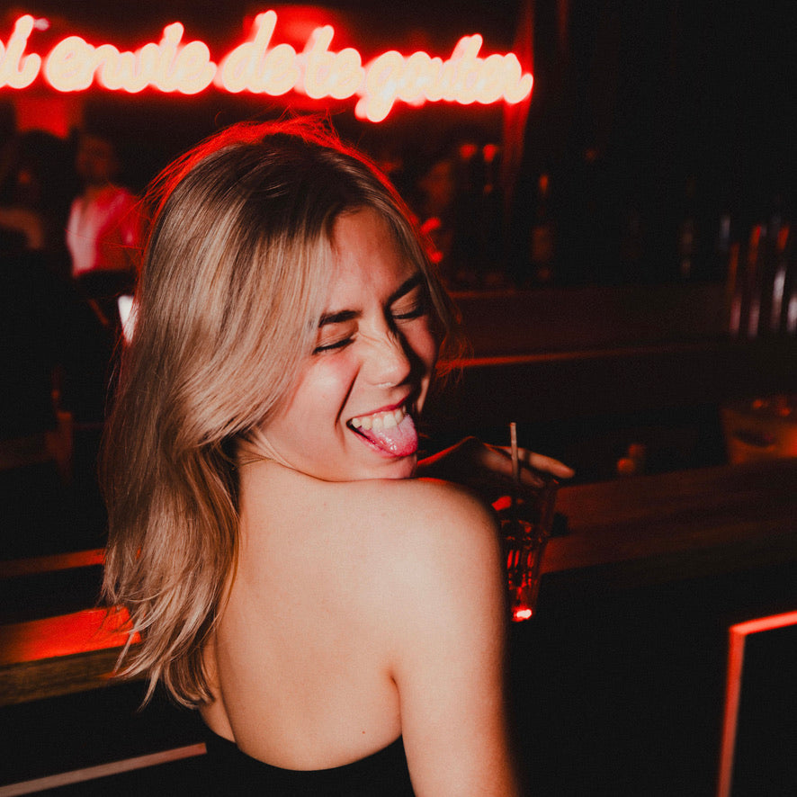 Portrait d'une femme blonde souriante et décontractée lors d'une soirée Crush Party à Paris. Capturée dans une ambiance festive avec une lumière tamisée et une enseigne néon rouge en arrière-plan, cette image reflète l'énergie conviviale et authentique des événements Crush Party, dédiés aux célibataires recherchant des rencontres uniques