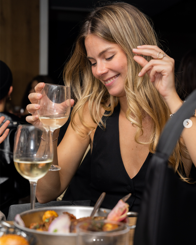 Jeune femme souriante en robe noire tenant un verre de vin blanc lors d'une soirée Crush Party dans un restaurant. Ambiance festive et conviviale avec des plats gastronomiques et des invités en arrière-plan, illustrant une rencontre authentique et dynamique.