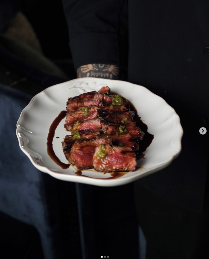 Assiette de viande grillée saignante avec une sauce et une garniture verte, servie par un serveur vêtu de noir avec un tatouage visible au poignet. Plat raffiné proposé lors des dîners Crush Party dans des restaurants partenaires comme Bazurto Paris.